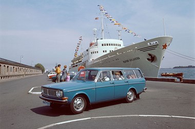 Bilde av en blå Volvo 145, parkert på en kai ved en stor båt. 