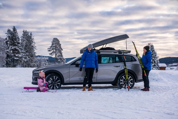 En familie med mor, far og barn står foran en Volvo med skiboks på taket. 